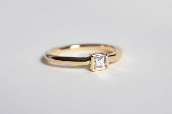 A yellow gold band that is rounded on one end and transitions to be squared on the other with a square framed white diamond setting