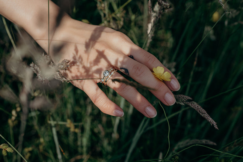 A hand in a grassy field modeling the white diamond crown kaori and white diamond enzo band stacked together on the ring finger. The hand is lightly grabbing a small yellow flower. 