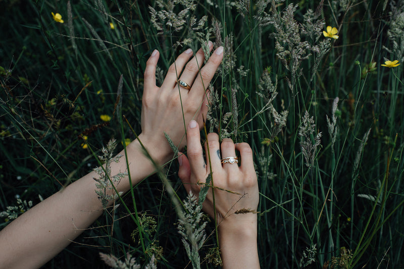 Two hands against a tall grassy field. The righ thand is modeling the whit diamond enzo band and white diamond crown kaori on the ring finger while the left hand has the green sapphire classic risa ring on the middle finger.   