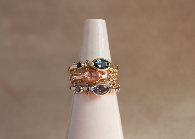 A close up view of a ring stack featuring the dark teal sapphire Kaori ring in yellow gold, the rose gold sunstone oval Kaori, and a violet montana sapphire Kaori in 14k white gold against a tan background.