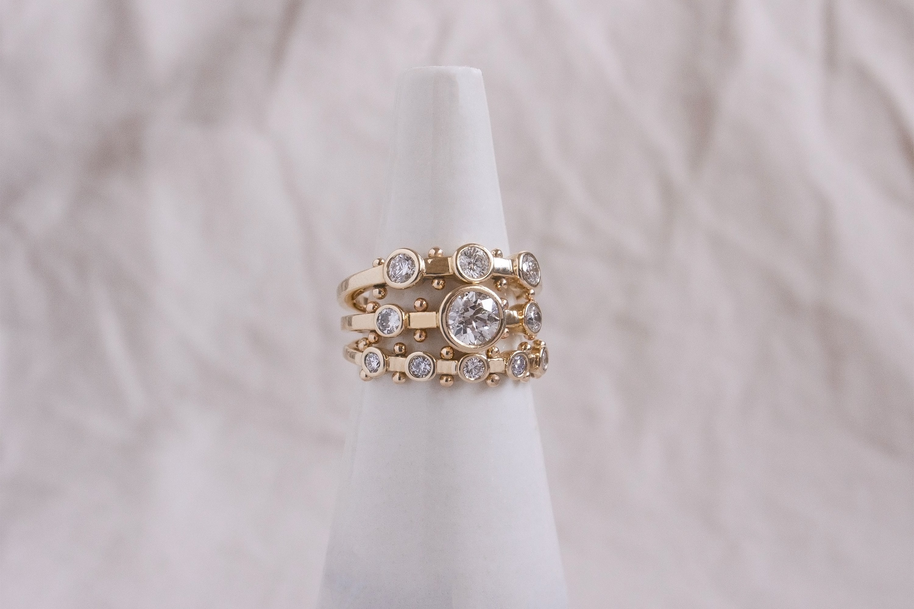 A close up view of a ring stack featuring the 3-stone, Crown Kaori, and 5-stone white diamond Koemi rings all with gold balls between each diamond setting along the sides of the band against a white background. 