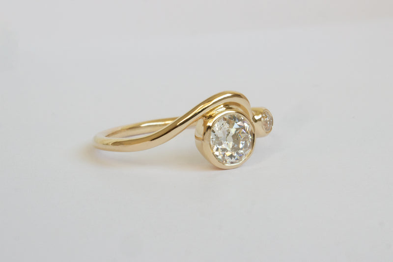 Side view of a bright white large round diamond and smaller diamond each framed in circular yellow gold setting on a wavy yellow gold band layered with another simple wavy yellow gold ring band