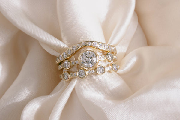 M. Hisae Crown Theia Ring, White Diamond Lana Band, and White Diamond 4-Stone Theia Ring stacked together on beautiful cream silk.