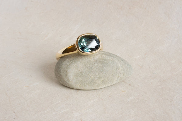 3.17ct cushion-cut green/blue sapphire ring in 14k yellow gold perched on a small stone.