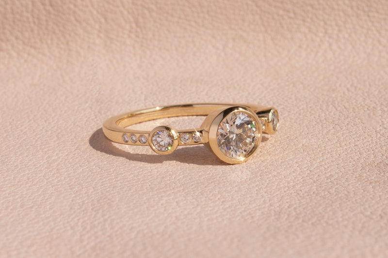M. Hisae three stone ring with a 0.5 carat center stone, two smaller side stones, and small diamonds flush set into band.